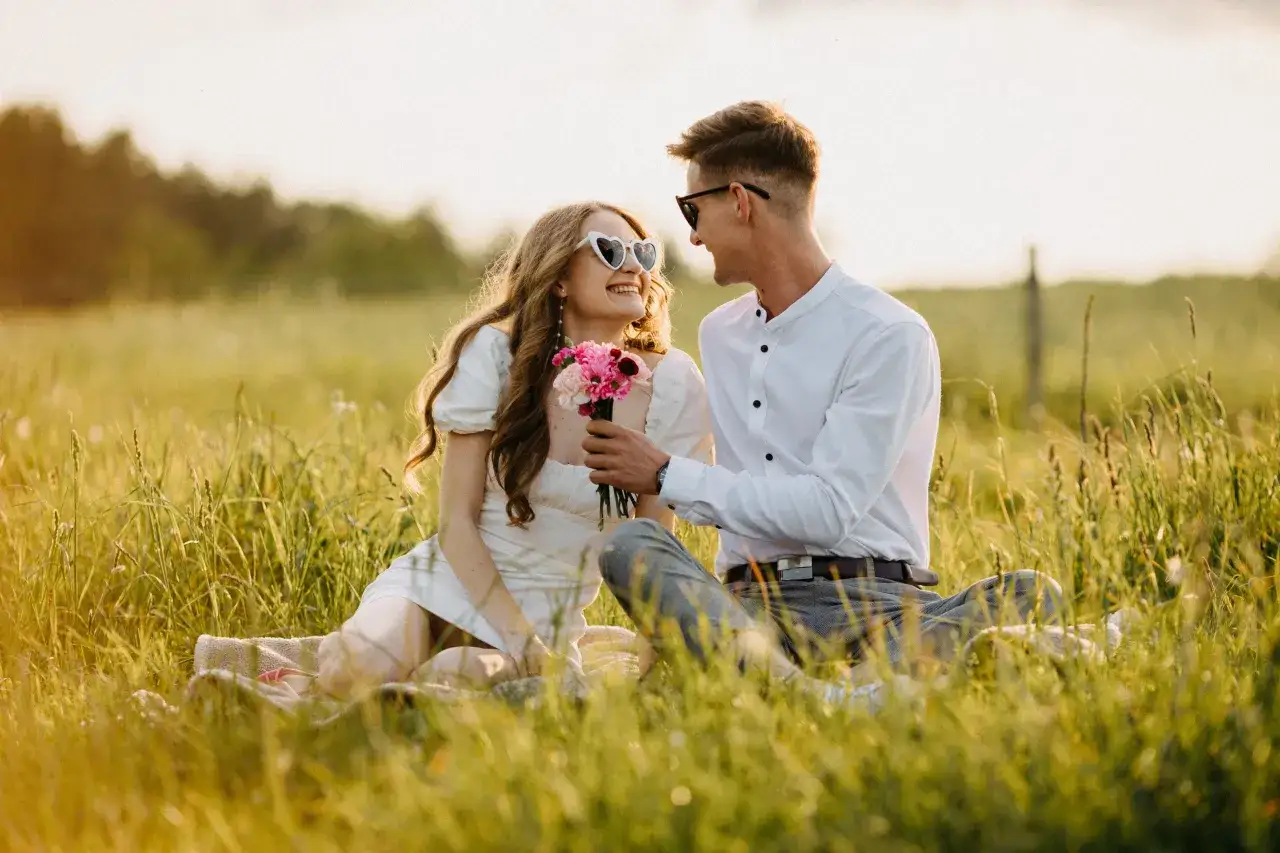 Uśmiechnięta para podczas sesji zdjęciowej dla par na łące. On daje jej bukiet różowych kwiatów.