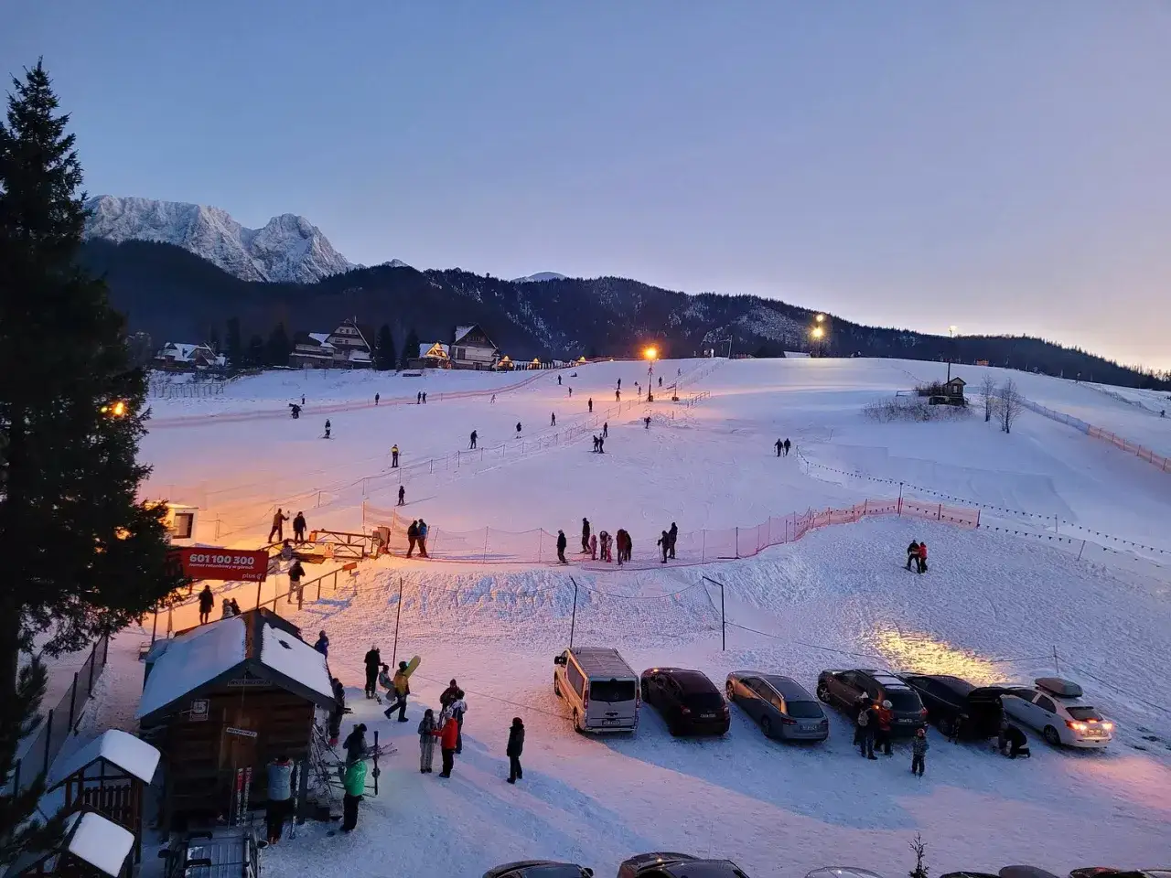 Oświetlony stok narciarski Budzowski Wierch wieczorem, z narciarzami i snowboardzistami, g&oacute;rami w tle i parkingiem.