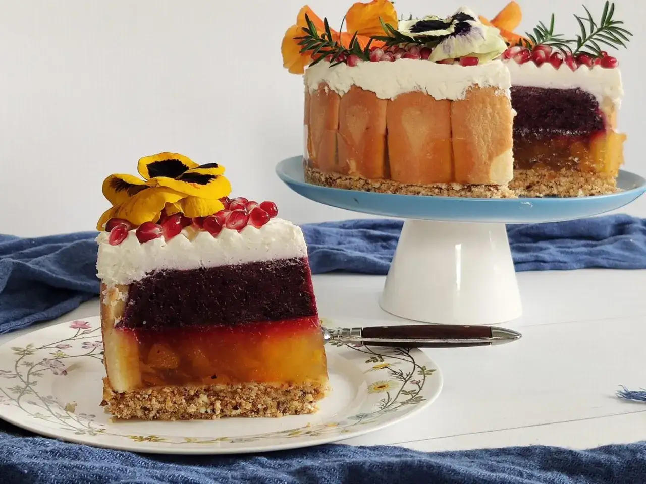 Kolorowe ciasto z biszkoptami, przełożone owocami i kremem, ozdobione kwiatami i granatem. Wygląda jak deser z bajki!