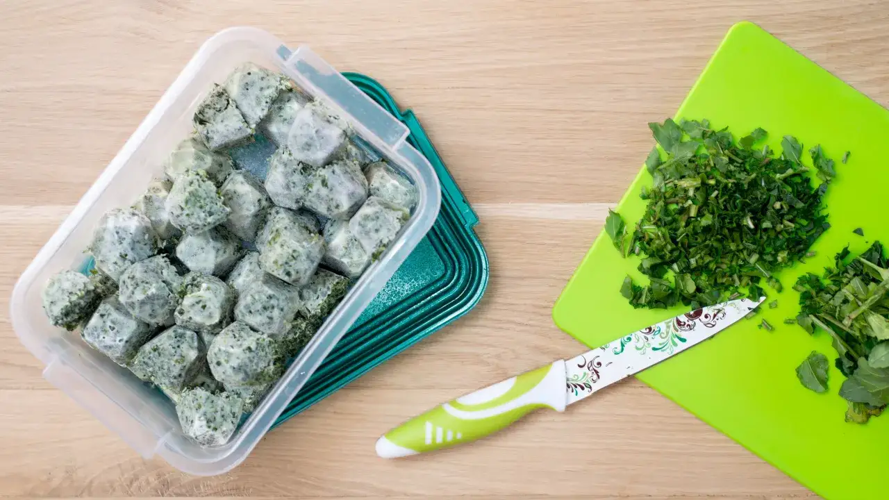 Kostki zamrożonej pietruszki w pojemniku, obok posiekana zielenina na desce i n&oacute;ż. Tak wygląda prosty spos&oacute;b, jak mrozić pietruszkę.