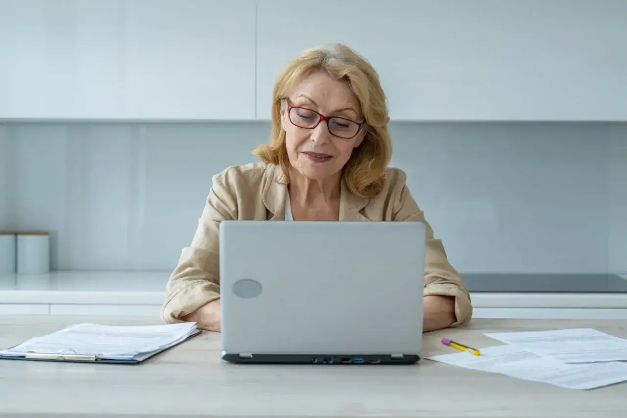 Kobieta po 50-tce szuka pracy, pokonując ageizm rekruterów. Analizuje dokumenty przy laptopie, przygotowując się do wyzwań rynku.