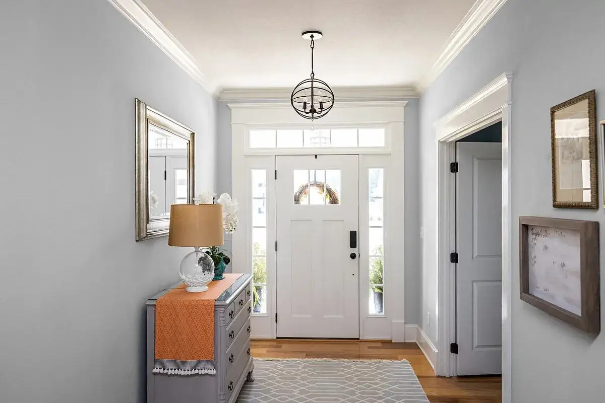 Jasny przedpok&oacute;j z białymi drzwiami i oknami. Po lewej komoda z lampą i lustrem, po prawej drzwi. Praktyczne ściany w przedpokoju odbijają światło.