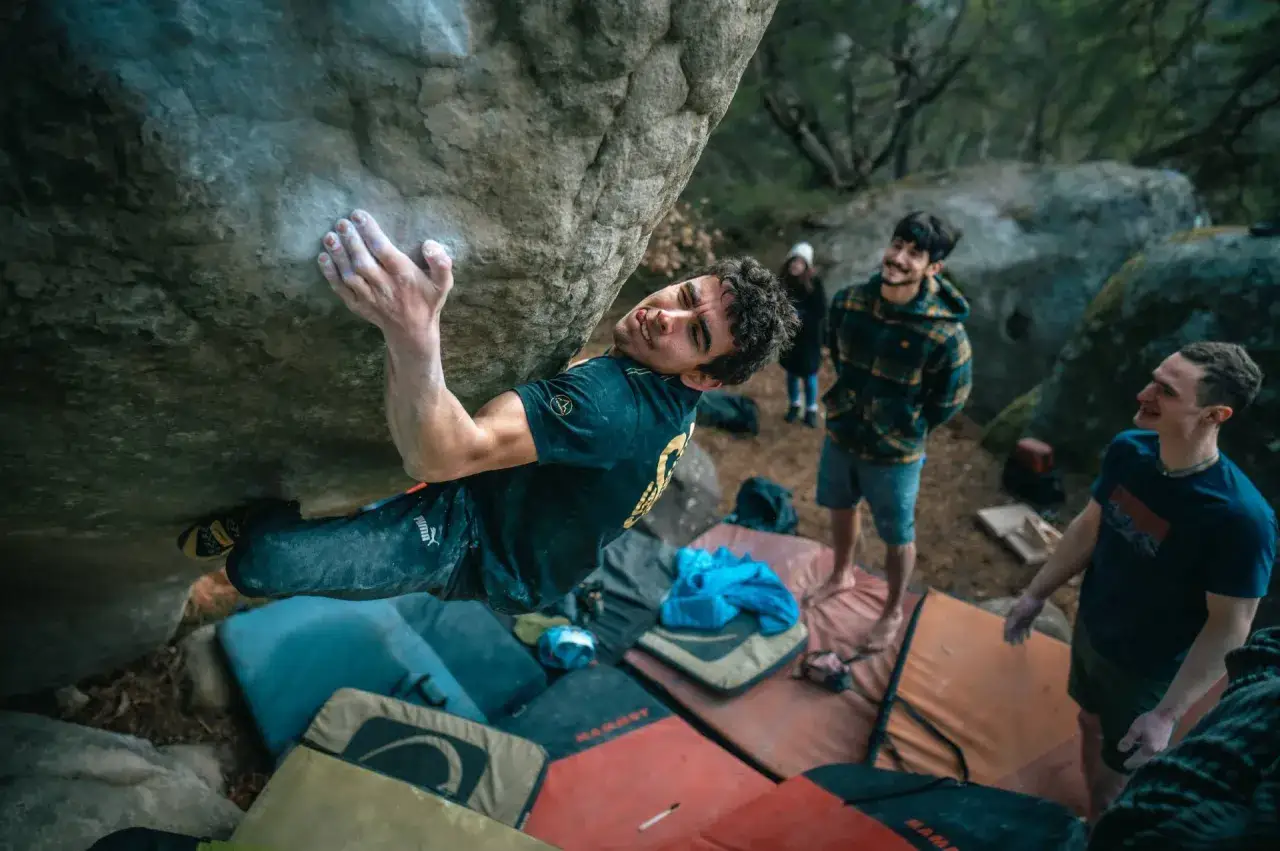 Wspinacz na skale podczas sesji **Fontainebleau bouldering**. W tle przyjaciele obserwują jego zmagania.
