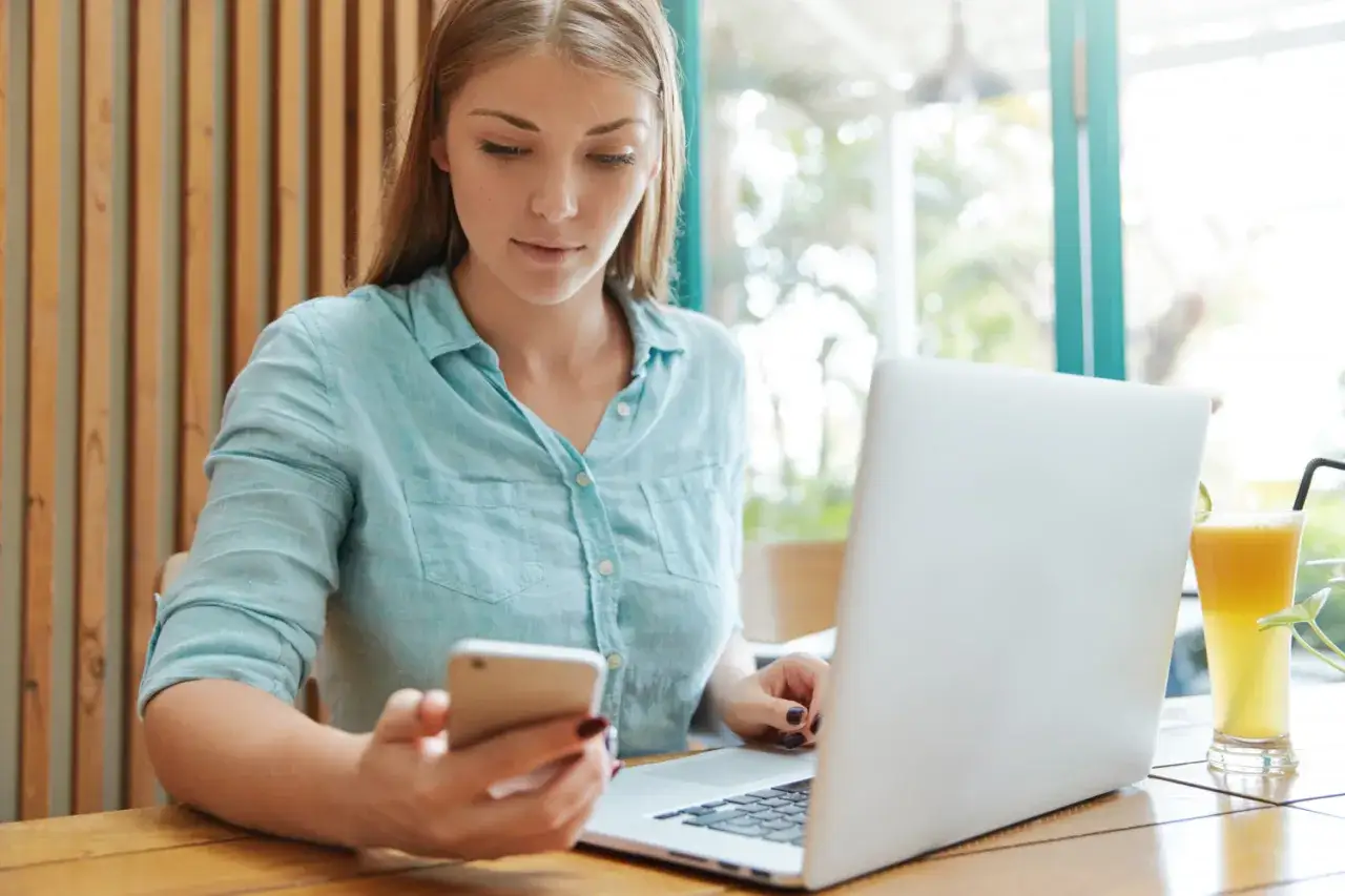 Kobieta sprawdza na telefonie ofertę kredytu z poręczycielem online, siedząc przy laptopie w kawiarni.