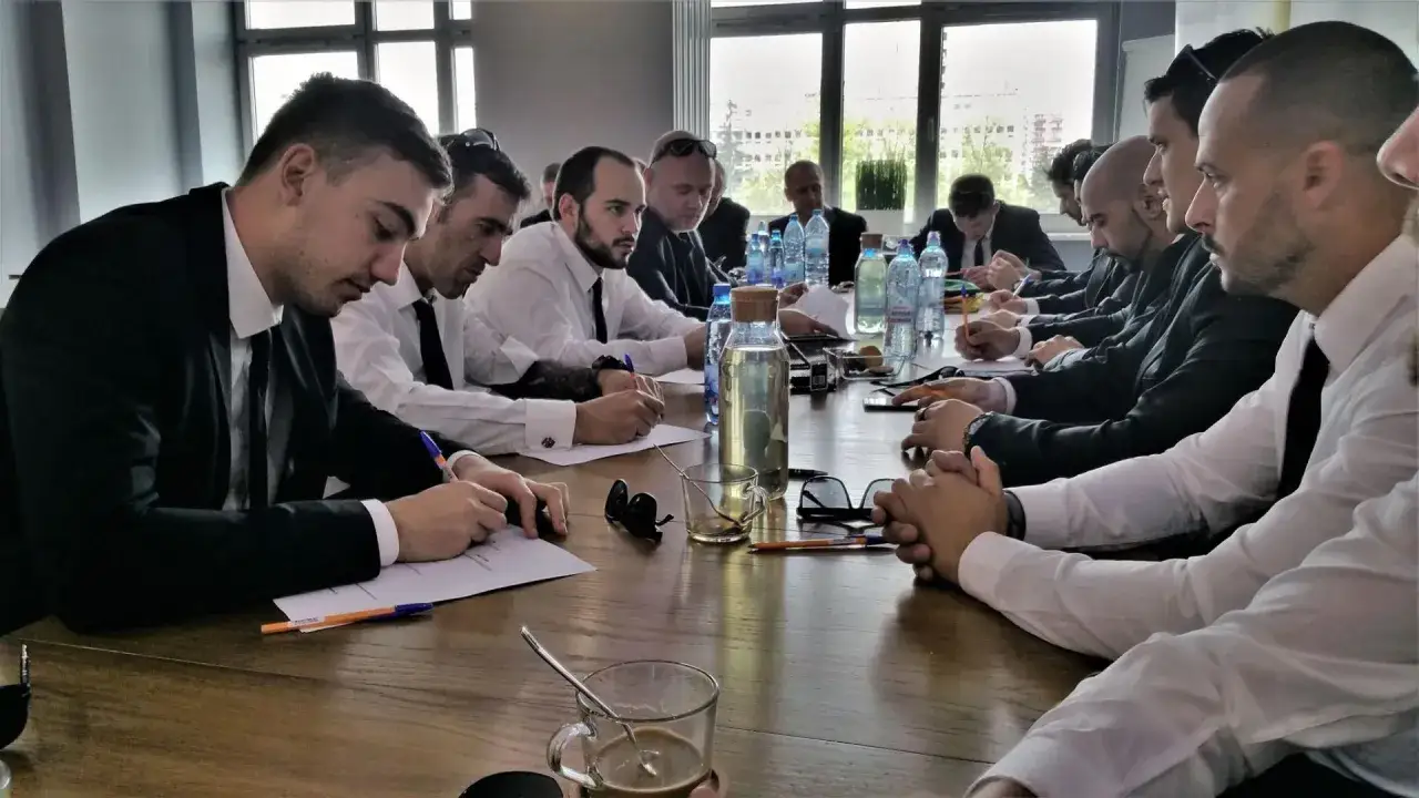 Uczestnicy kursu na ochroniarza skupieni przy stole, piszą notatki. W tle okna i butelki z wodą.