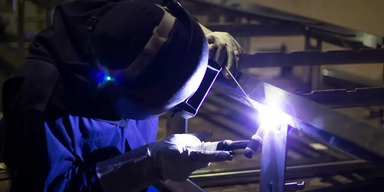 Spawanie aluminium: pracownik w masce spawalniczej i rękawicach precyzyjnie łączy metalowe elementy, tworząc jasny łuk świetlny.