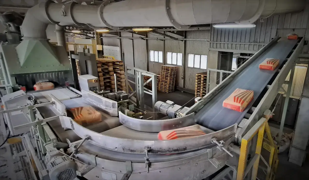Taśmociąg transportuje worki z materiałem, z którego się robi cement. W tle widać palety i elementy linii produkcyjnej.