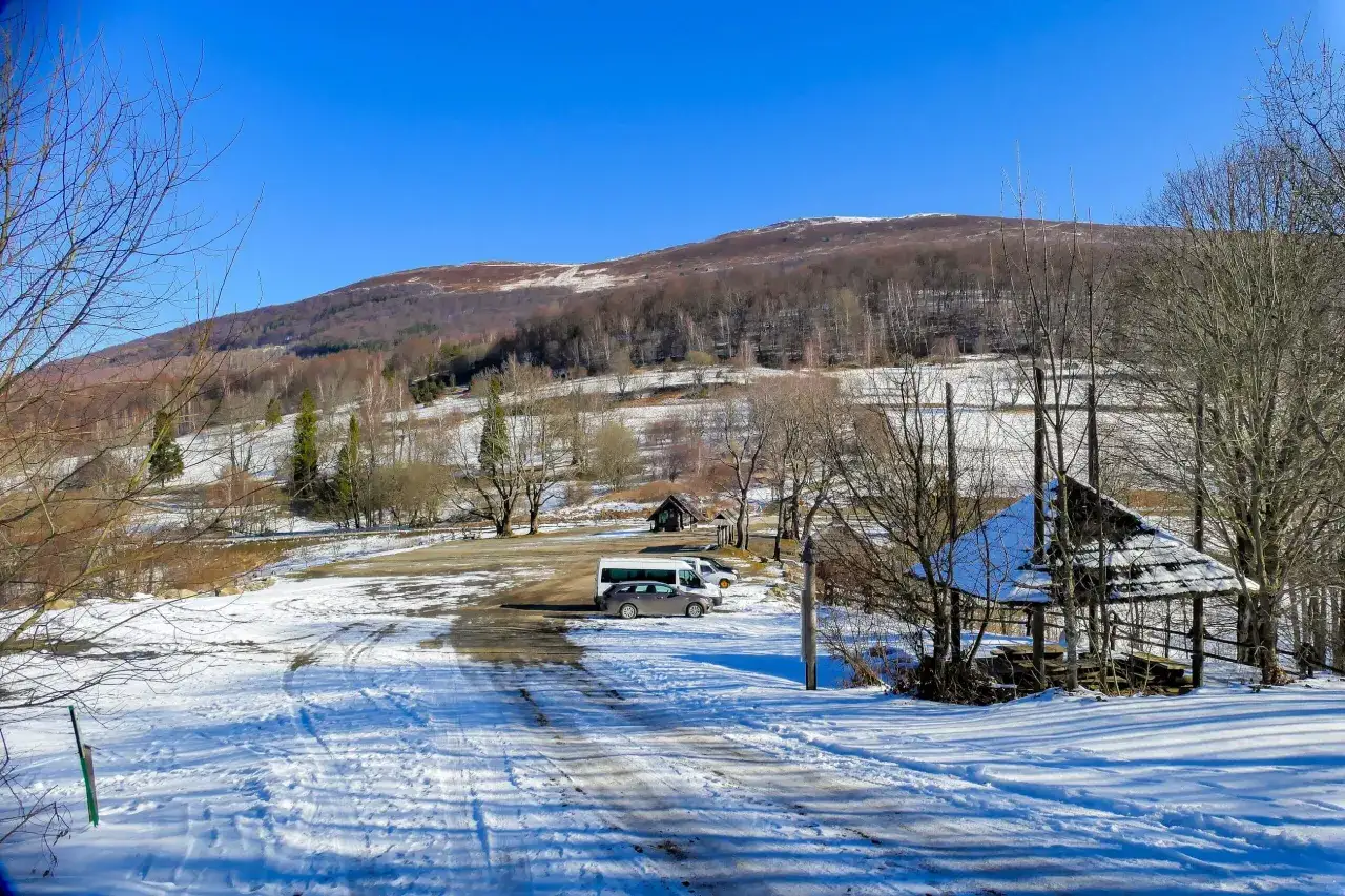 Zimowy krajobraz z bac&oacute;wką pod Małą Rawką. Na śniegu widać ślady opon i ścieżki. W tle g&oacute;ra porośnięta lasem.