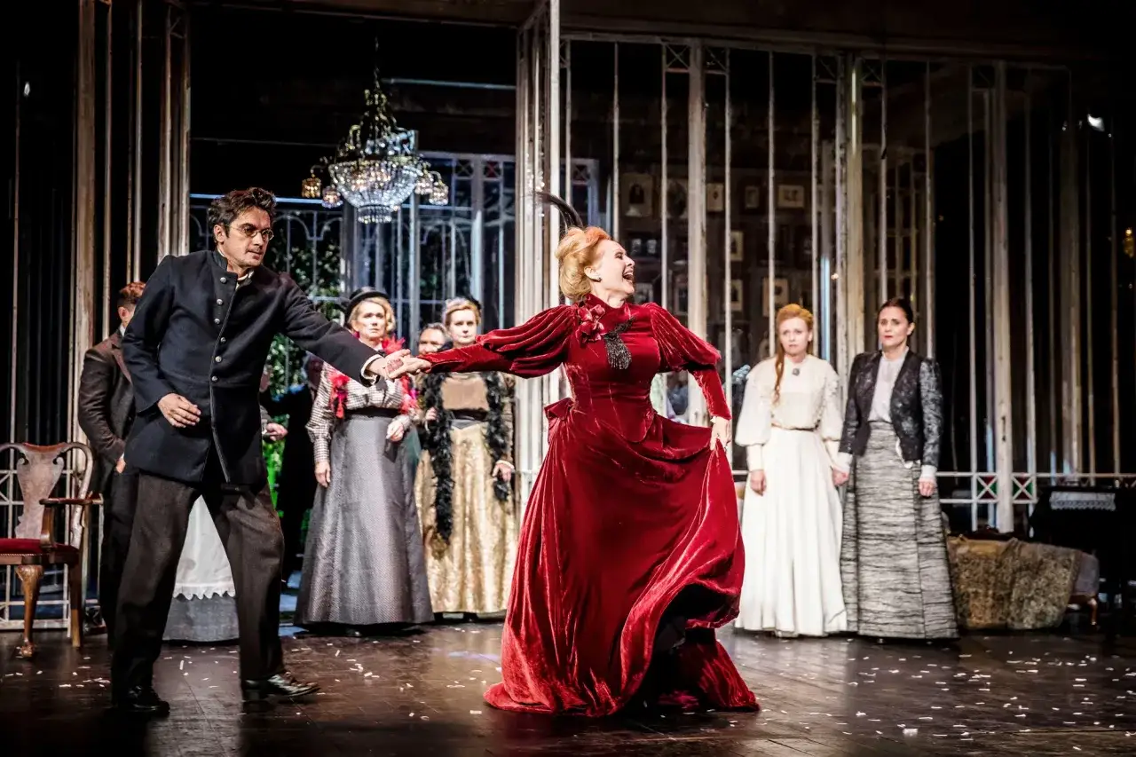 Aktorzy na scenie, w centrum kobieta w czerwonej sukni, tworzą wiśniowy sad spektakl.