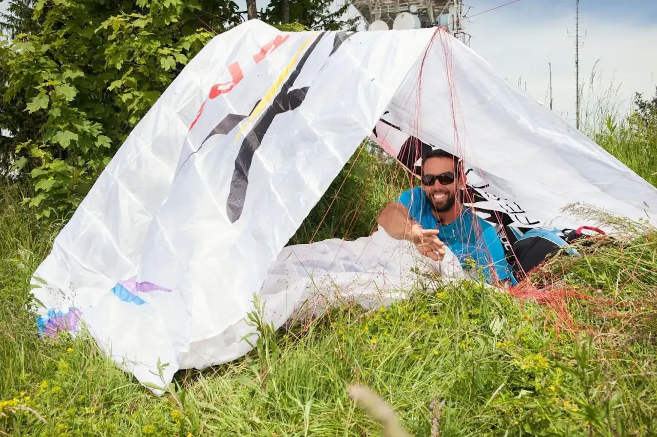 Uśmiechnięty mężczyzna w okularach przeciwsłonecznych leży pod białym materiałem, kt&oacute;ry wygląda jak rozłożony spadochron lub namiot. Pokazuje, jak rozłożyć namiot.