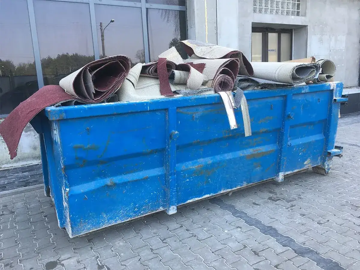 Niebieski kontener na śmieci w Warszawie wypełniony zrolowanymi dywanami. Zastanawiasz się, ile kosztuje kontener na śmieci Warszawa?