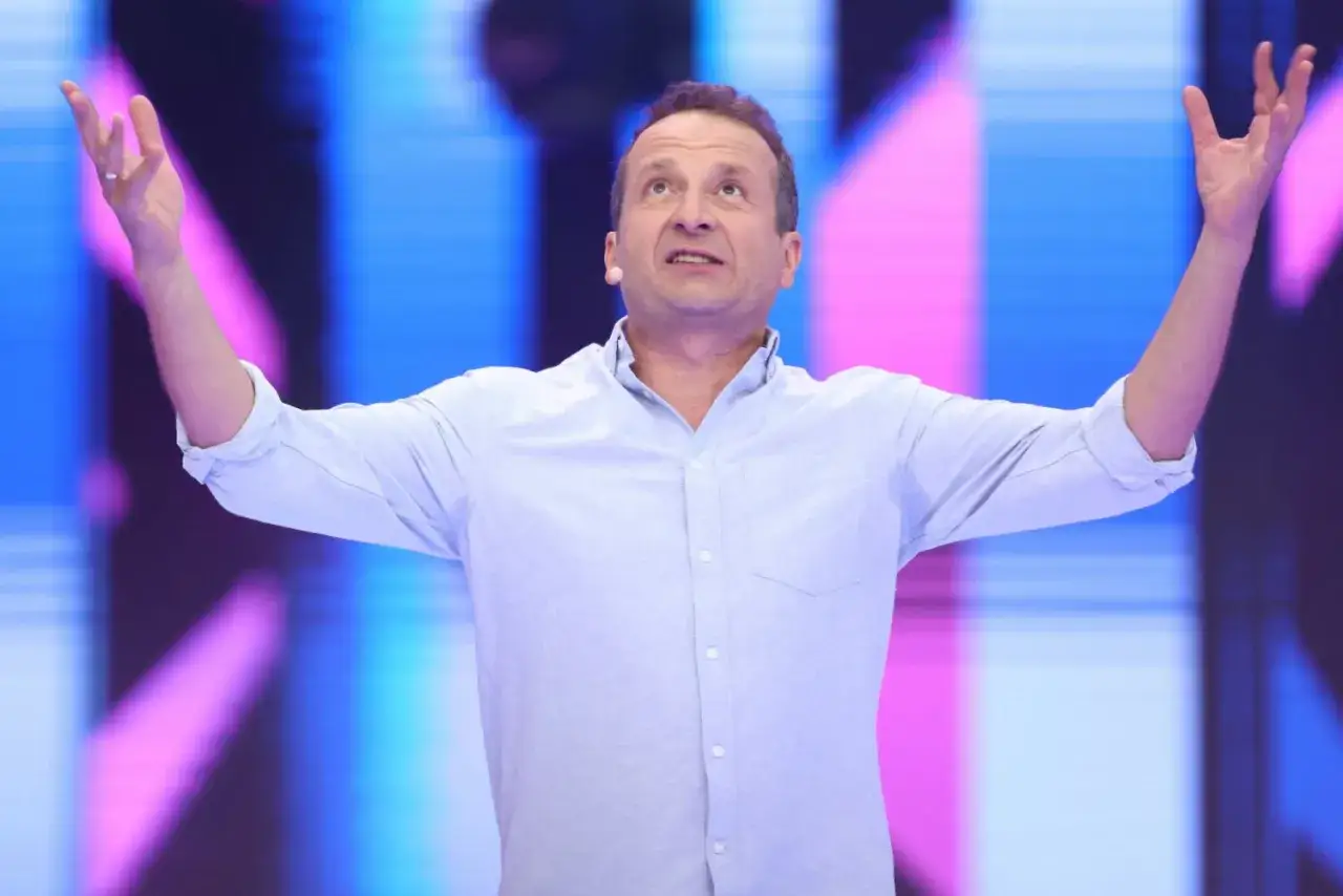 Aktor z Kabaretu Moralnego Niepokoju w błękitnej koszuli, z rękami rozłożonymi na boki, w trakcie jednego ze skeczy.