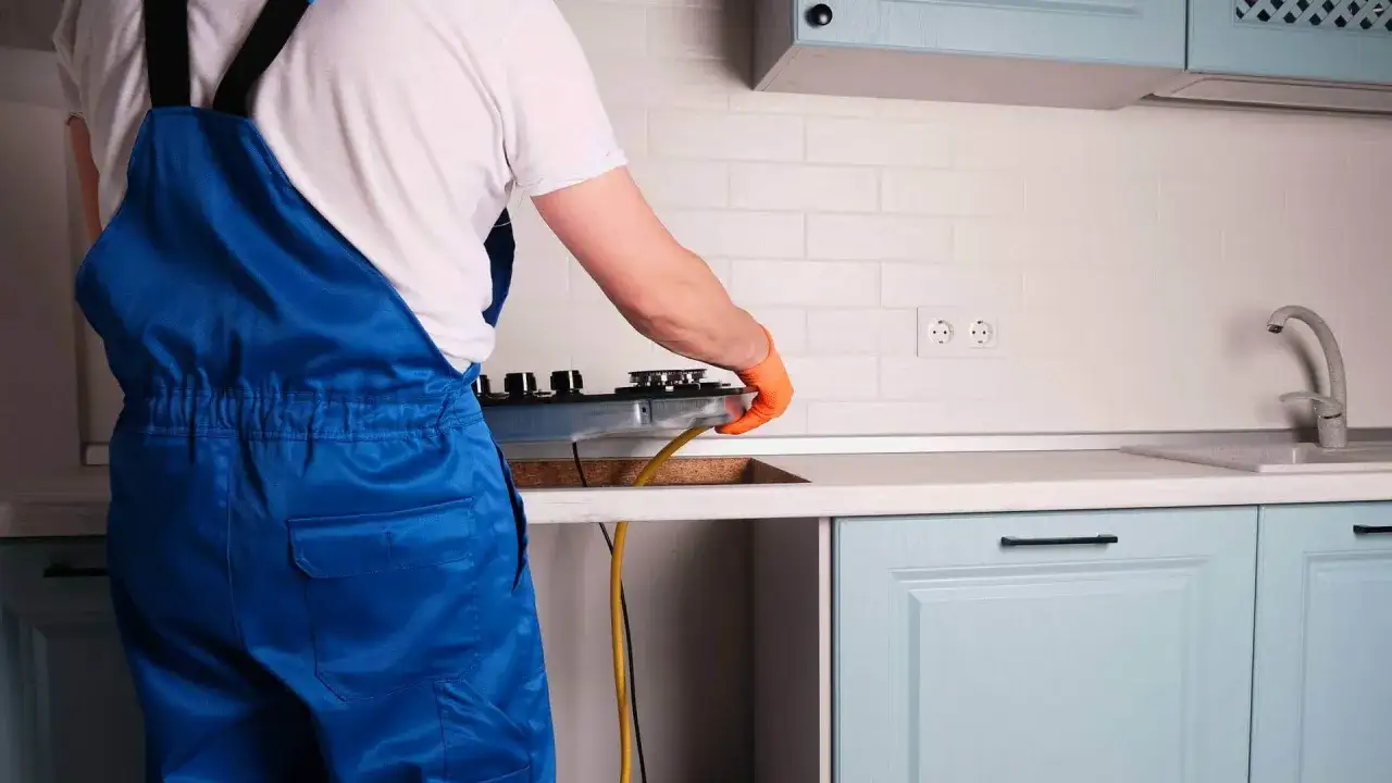 Fachowiec w niebieskim kombinezonie dokonuje podłączenia płyty gazowej w kuchni.