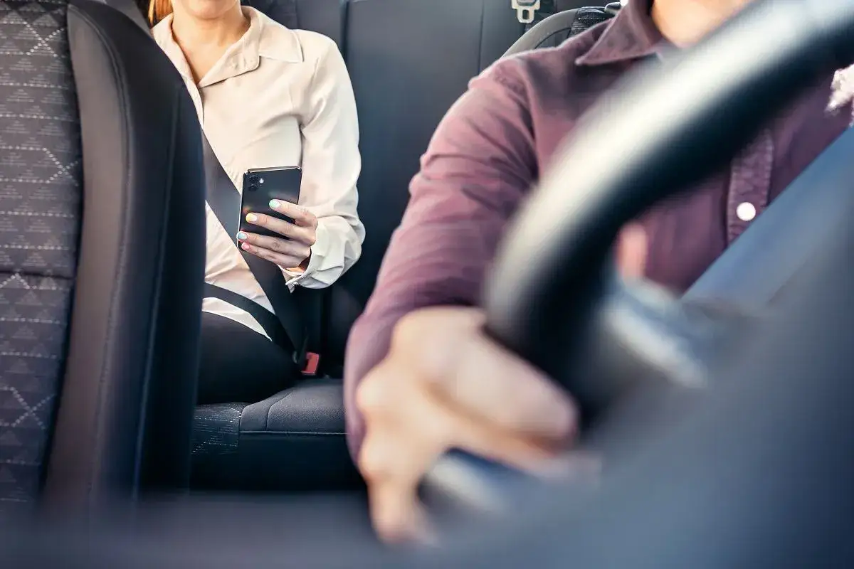Pasażerka w jasnej bluzce trzyma telefon, a kierowca w bordowej koszuli prowadzi auto. Czy taksówkarz musi zapinać pasy? Bezpieczeństwo przede wszystkim!