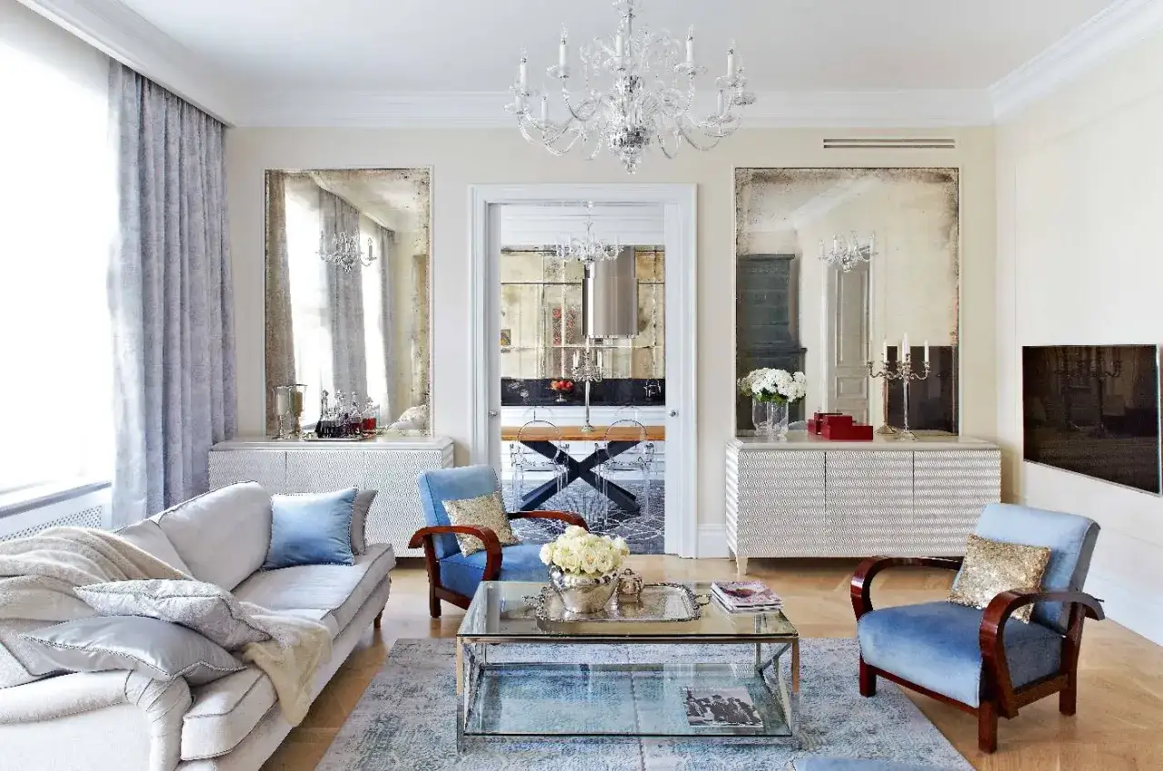 Elegancki salon z błękitnymi fotelami i sofą. W tle widać kuchnię, a na ścianach wiszą postarzone lustra, które dodają wnętrzu charakteru.