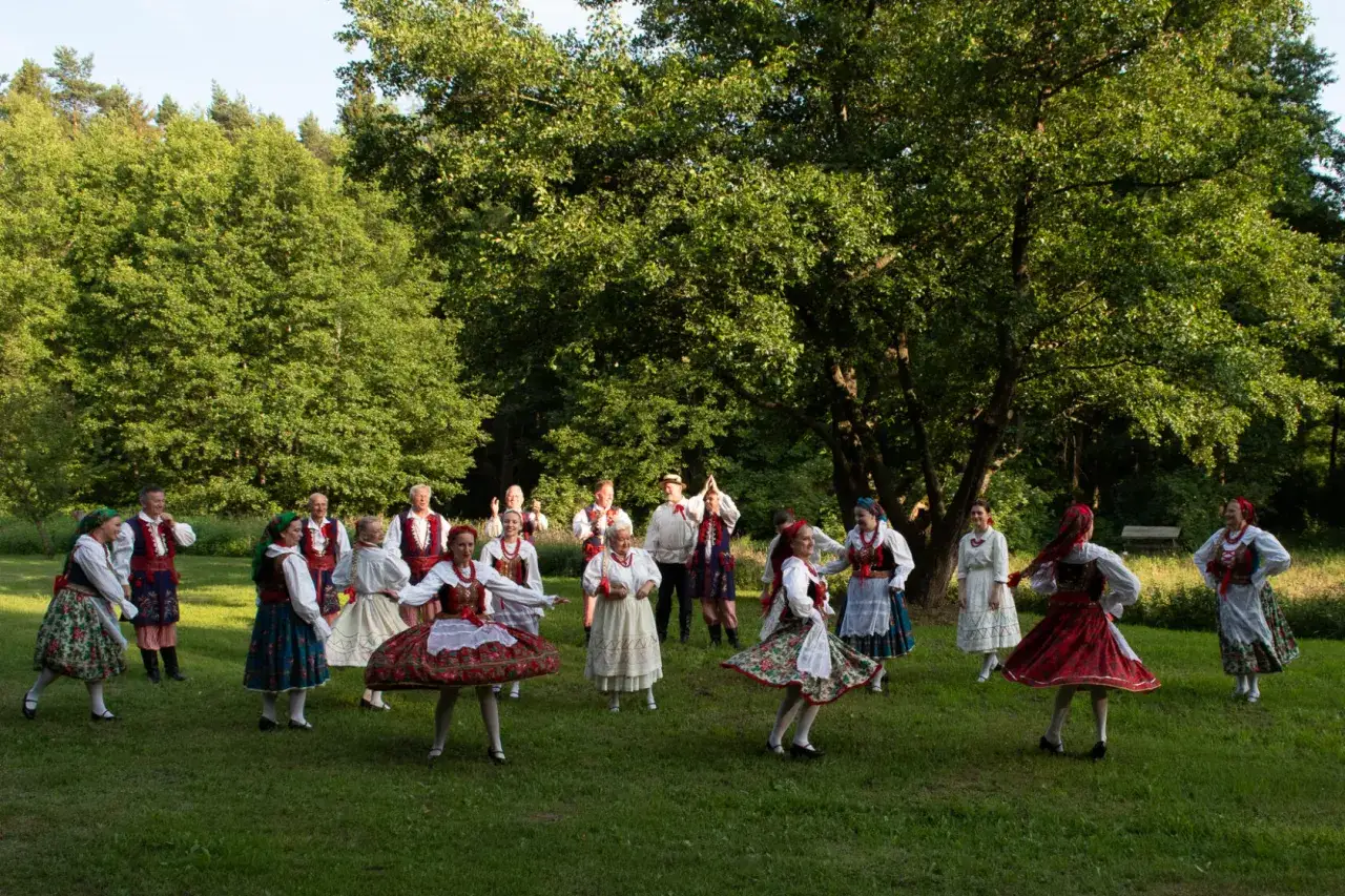 Grupa ludzi w tradycyjnych strojach ludowych tańczy na trawie. Słychać wiejskie przyśpiewki teksty, które niosą się po lesie.