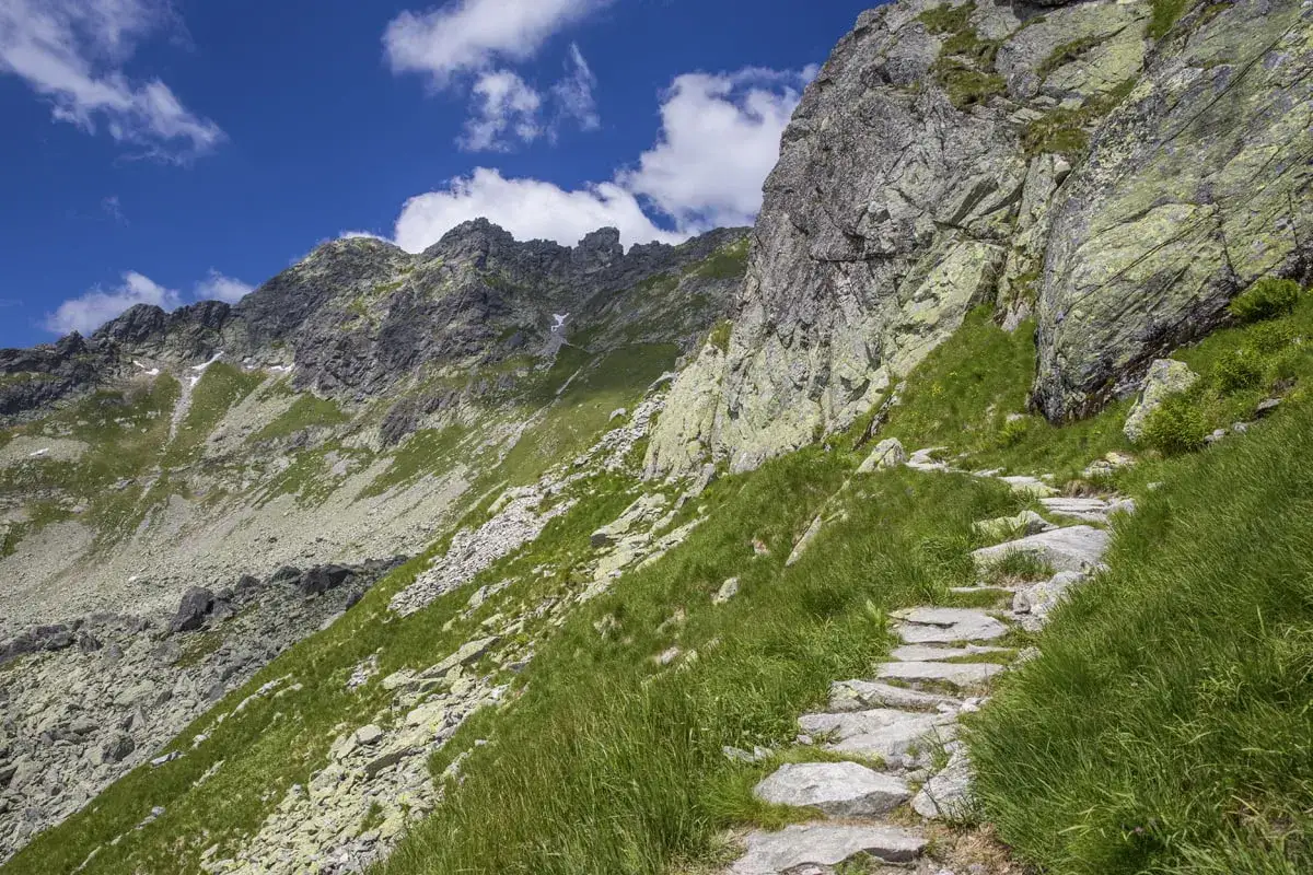 Kamienista ścieżka wije się po zboczu górskim, prowadząc w stronę skalistych szczytów. Zielona trawa otacza szlak na Zawrat, a błękitne niebo z chmurami dodaje uroku.