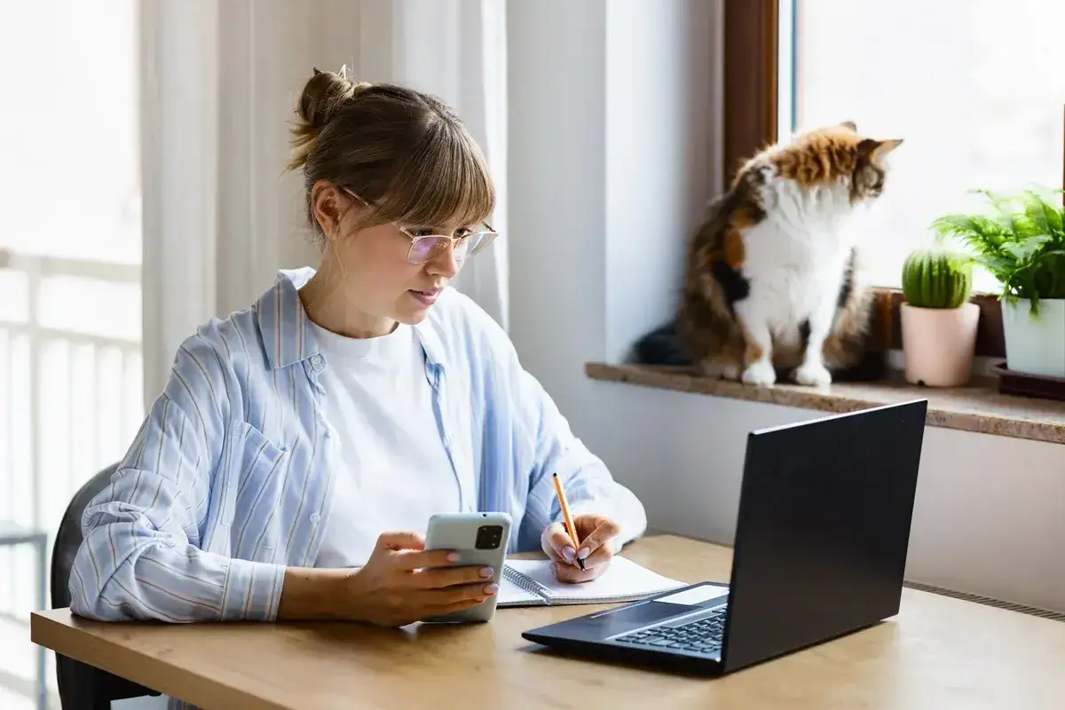 Młoda kobieta z kotem na parapecie pracuje przy laptopie, notując coś. To obrazek pokazujący, freelancer co to znaczy w praktyce.