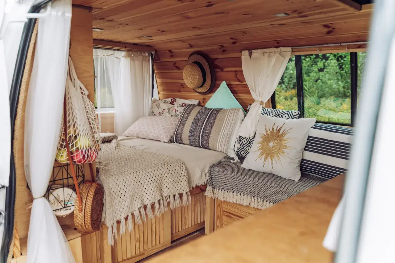 Przytulne wnętrze kampera z drewnianymi panelami, wygodnym ł&oacute;żkiem, mn&oacute;stwem poduszek i plecionym koszem. Idealne na podr&oacute;ż!