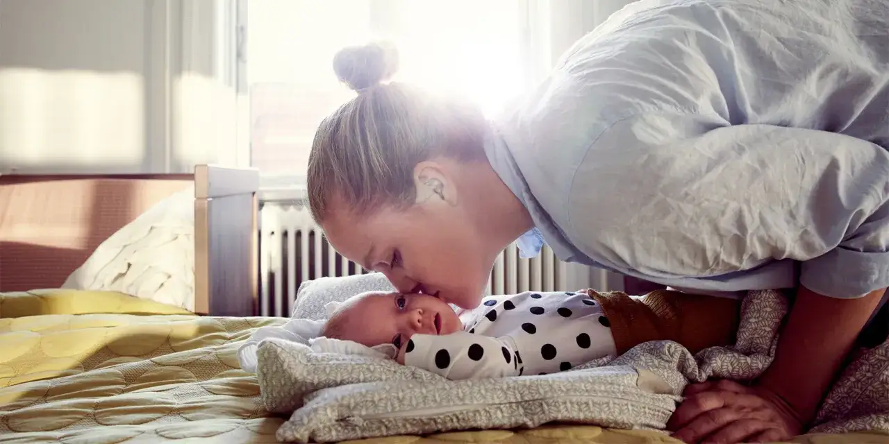 Mama całuje dziecko, które leży w łóżeczku. To piękny moment, być może pierwszy skok rozwojowy u noworodka.