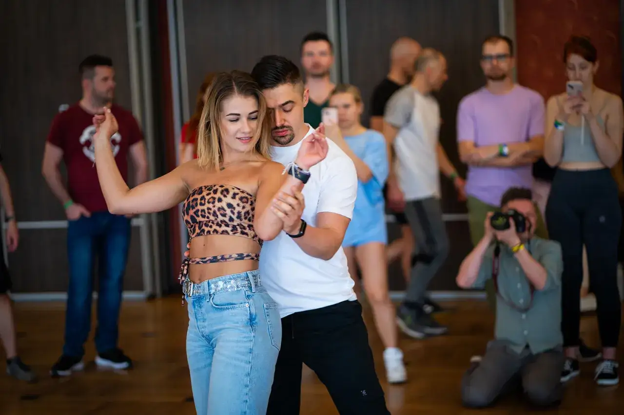 Para tańczy bachata, poznając nazwy krok&oacute;w. W tle inni uczestnicy i fotograf.