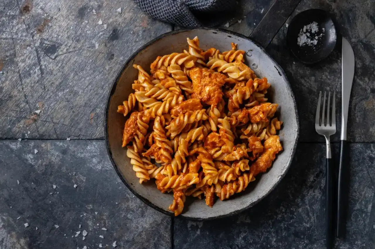 Fusilli z kurczakiem w sosie z czerwonego pesto ze słoika, podane w misce. Obok widelec i n&oacute;ż.