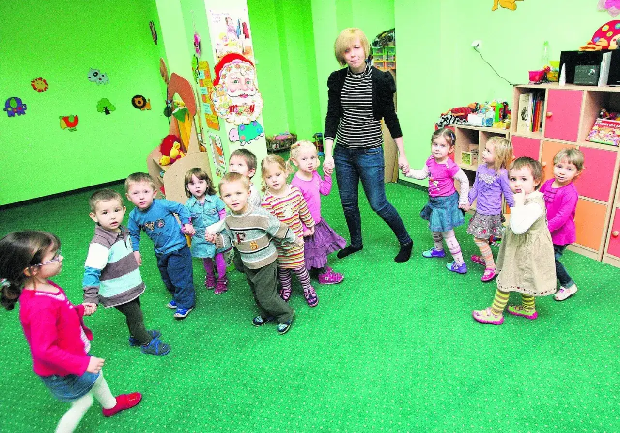 Dzieci trzymają się za ręce w żłobku, który podlega pod kuratorium. Nauczycielka prowadzi grupę.