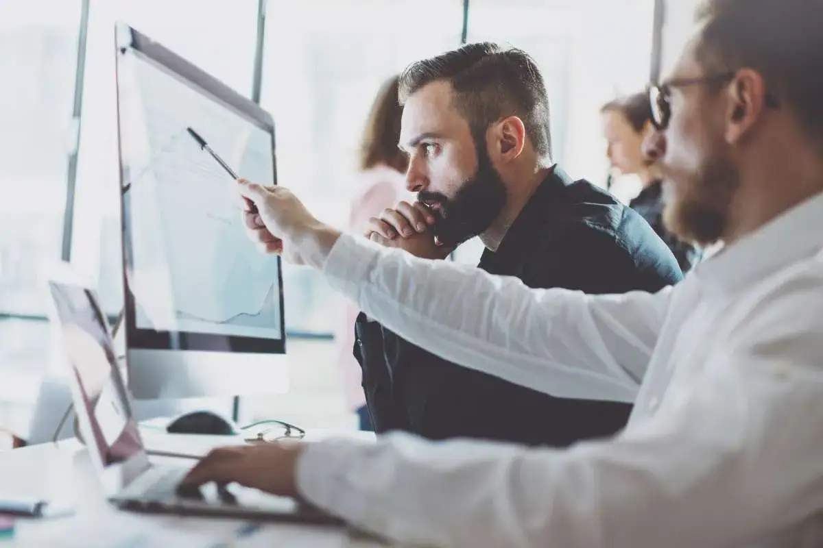 Dw&oacute;ch mężczyzn analizuje wykres na monitorze. Jeden z nich wskazuje pi&oacute;rem na dane, zastanawiając się 