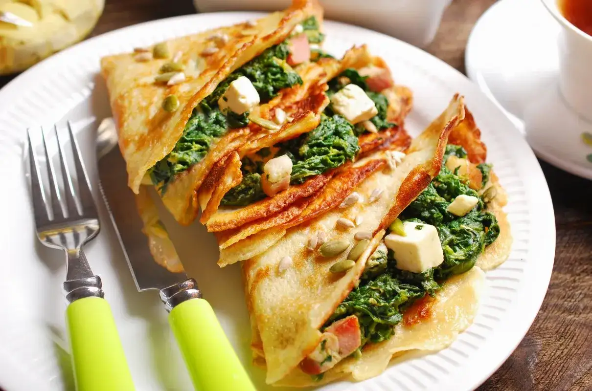 Naleśniki ze szpinakiem i serem feta na obiad, posypane pestkami. Obok widelec i nóż z zielonymi uchwytami.