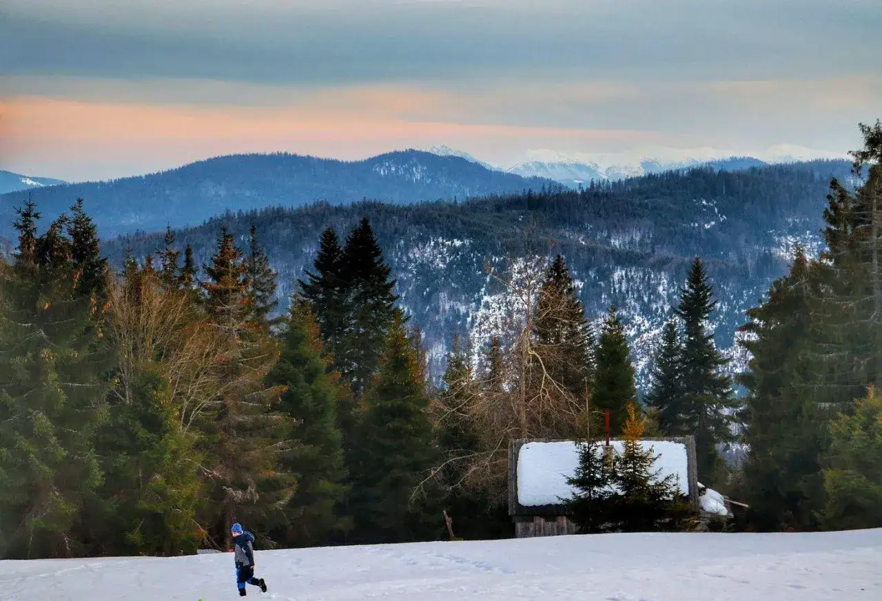 Dziecko biega po śniegu obok bac&oacute;wki Krawc&oacute;w Wierch. W tle g&oacute;ry i lasy.