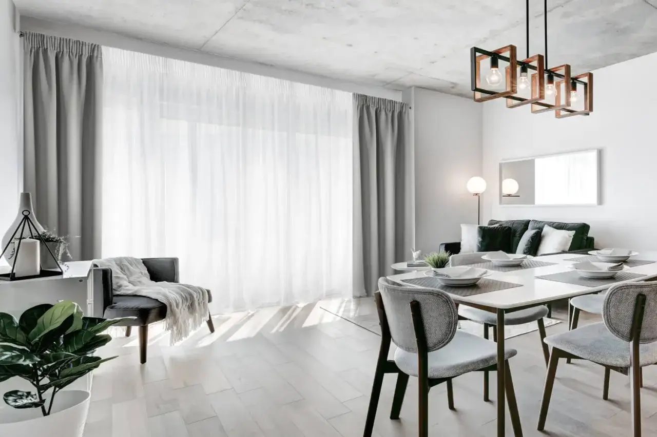 Jasne, nowoczesne firany w salonie tworzą przytulną atmosferę. Widać też stół z krzesłami, sofę i designerską lampę.