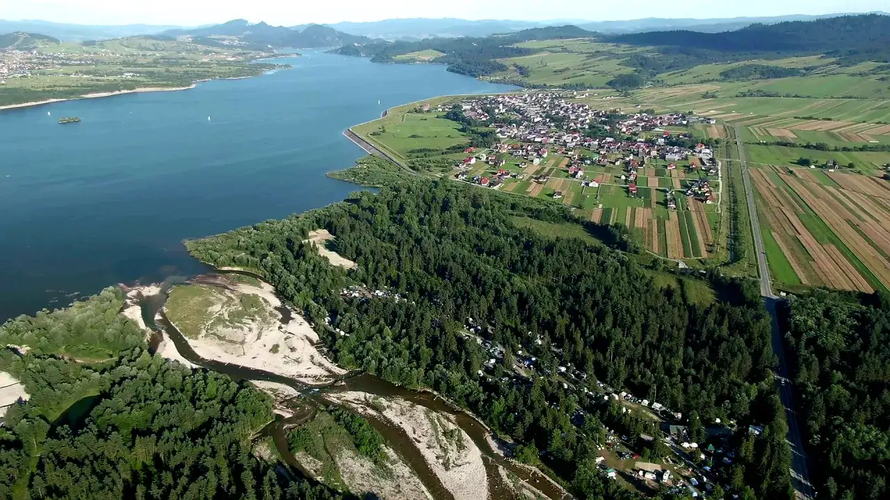 Malowniczy widok na jezioro, las i pola uprawne. Wśród drzew widać rozległy camping łęg frydman z namiotami i samochodami.