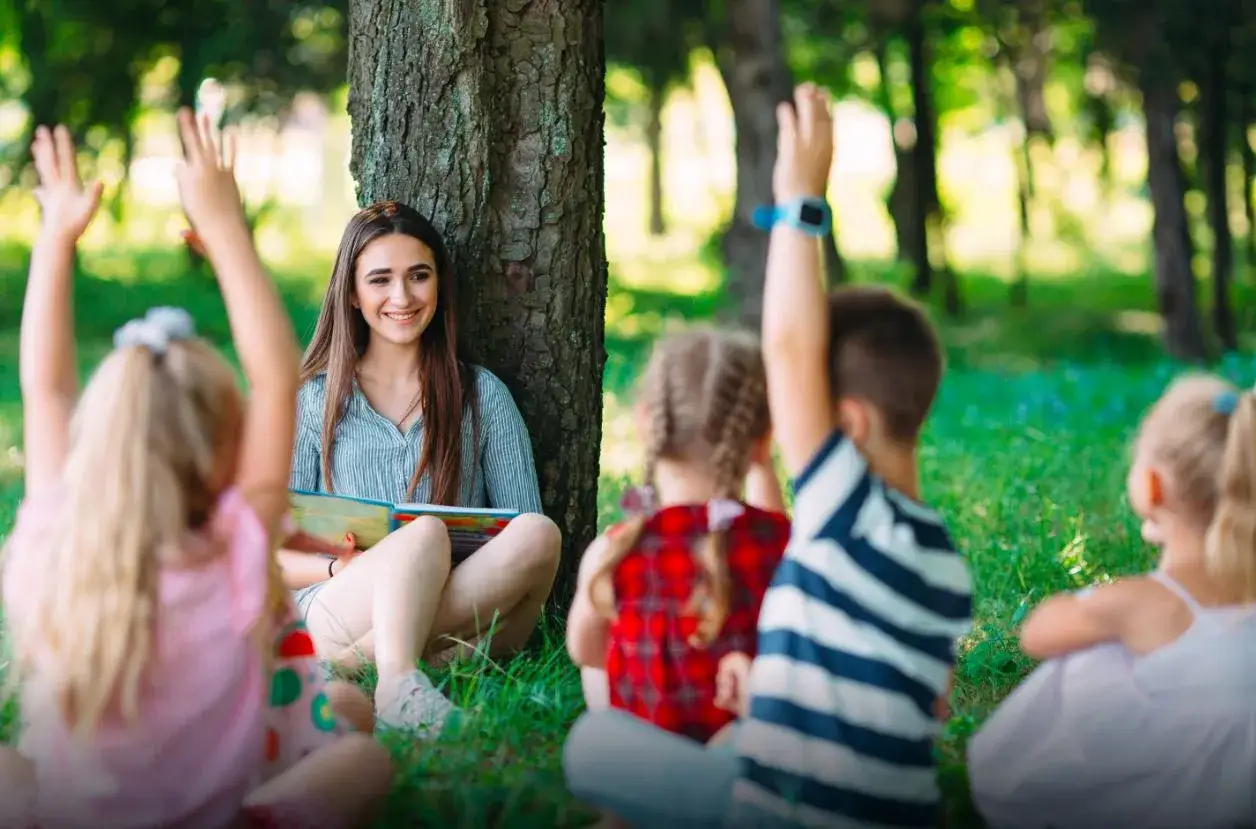 Uśmiechnięta młoda kobieta czyta książkę dzieciom na trawie. Widać, że rodzic może być opiekunem na wycieczce szkolnej.
