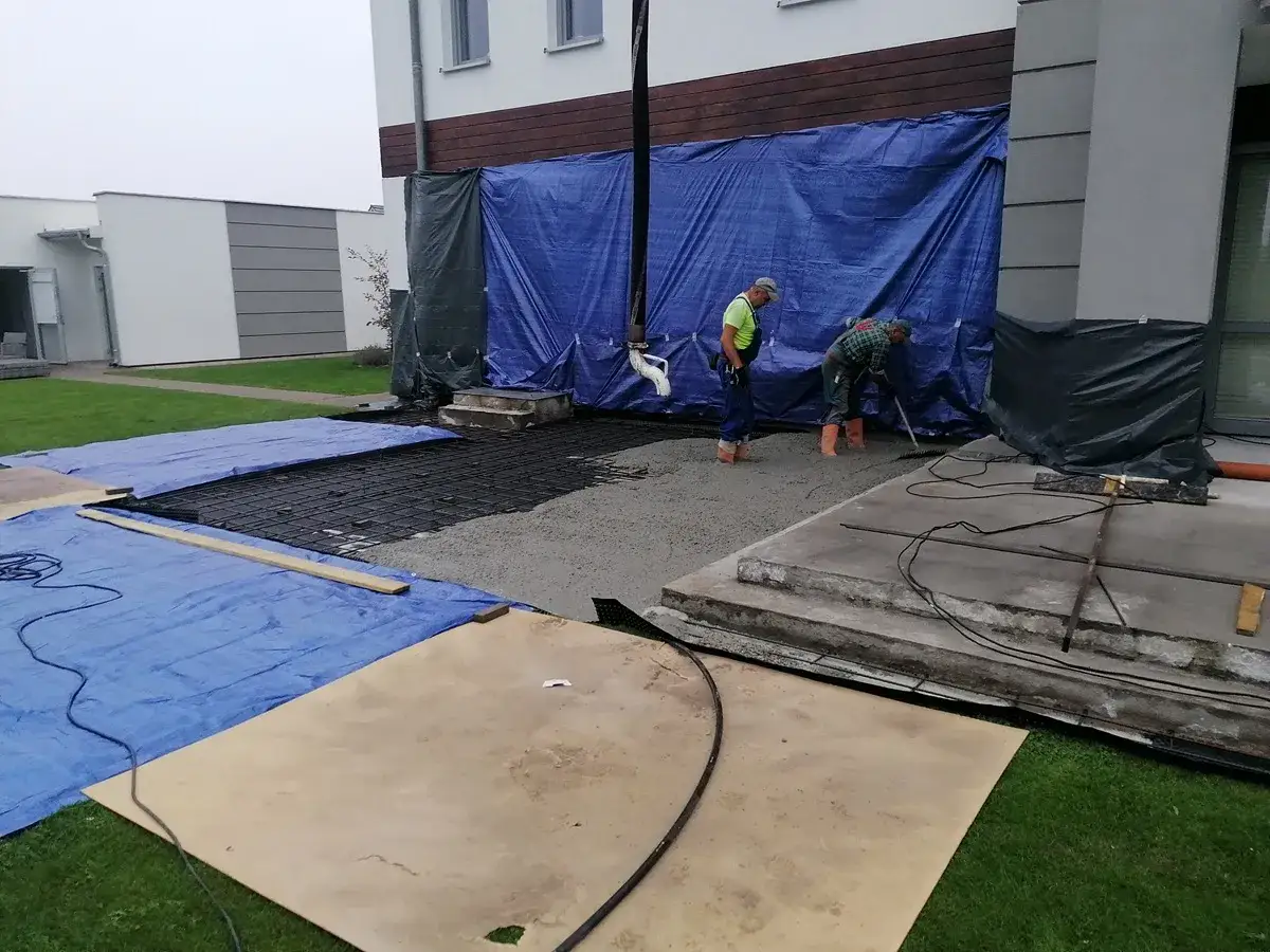 Pracownicy wykonują układanie płyt tarasowych na suchym betonie. Widać zbrojenie i świeży beton.