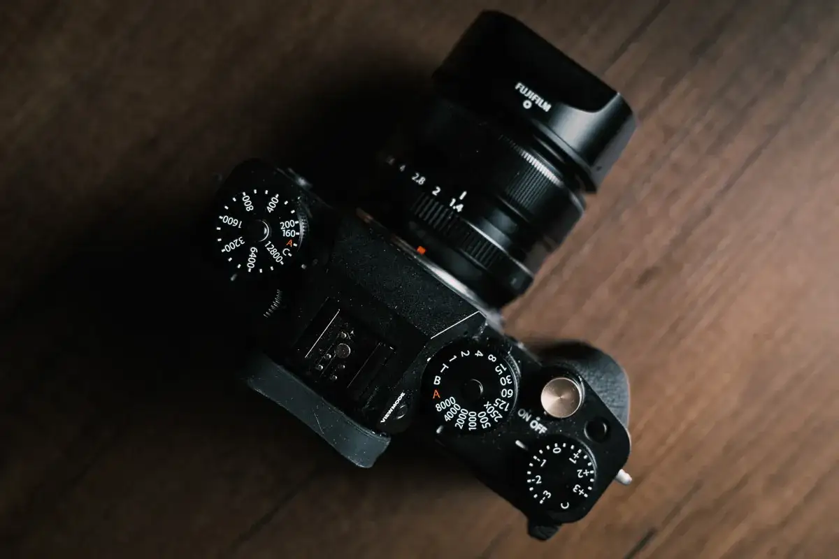 Czarny aparat Fujifilm X-T4 z obiektywem na drewnianym tle. Widoczne pokrętła ISO i przysłony.