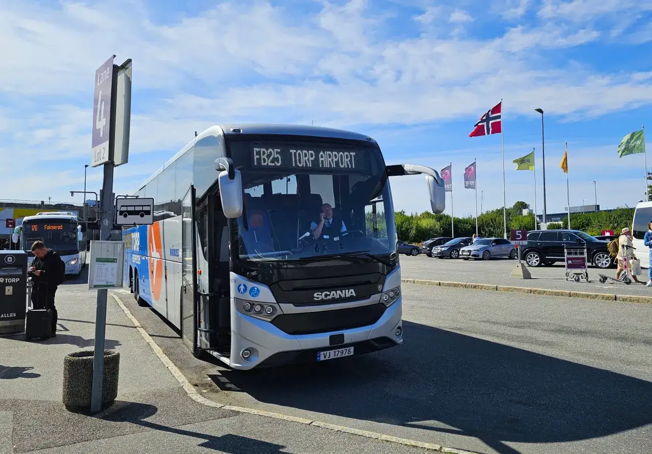 Autobus FB25 Torp Airport czeka na pasie 4, Lot 2. W tle widać inne pojazdy i flagi Norwegii.