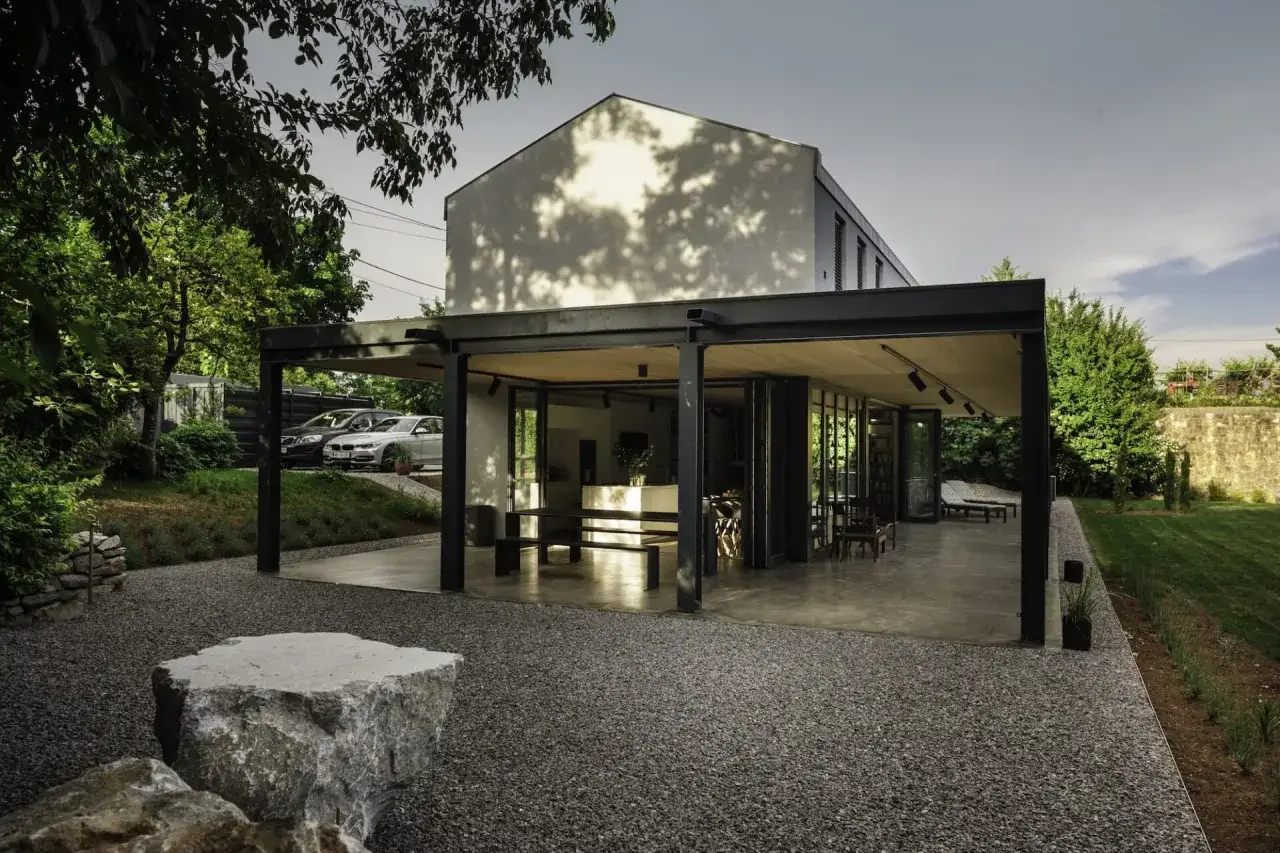 Nowoczesny projekt domu z tarasem, gdzie przeszklone ściany otwierają wnętrze na ogr&oacute;d. Dwa samochody zaparkowane obok.