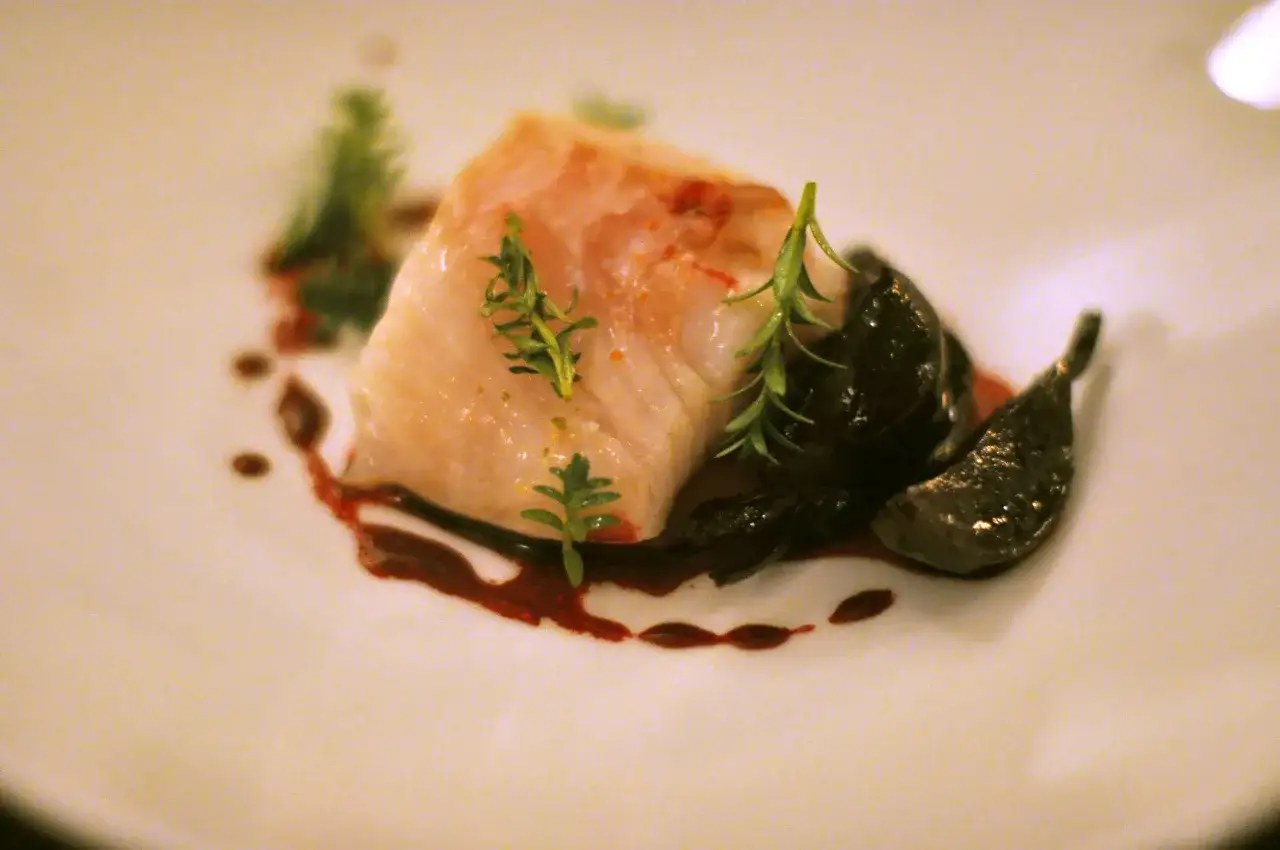 Delikatny kawałek ryby z ciemnymi, błyszczącymi dodatkami i zielonymi gałązkami. Czy ryba jest wegetariańska? To pytanie pozostaje bez odpowiedzi.