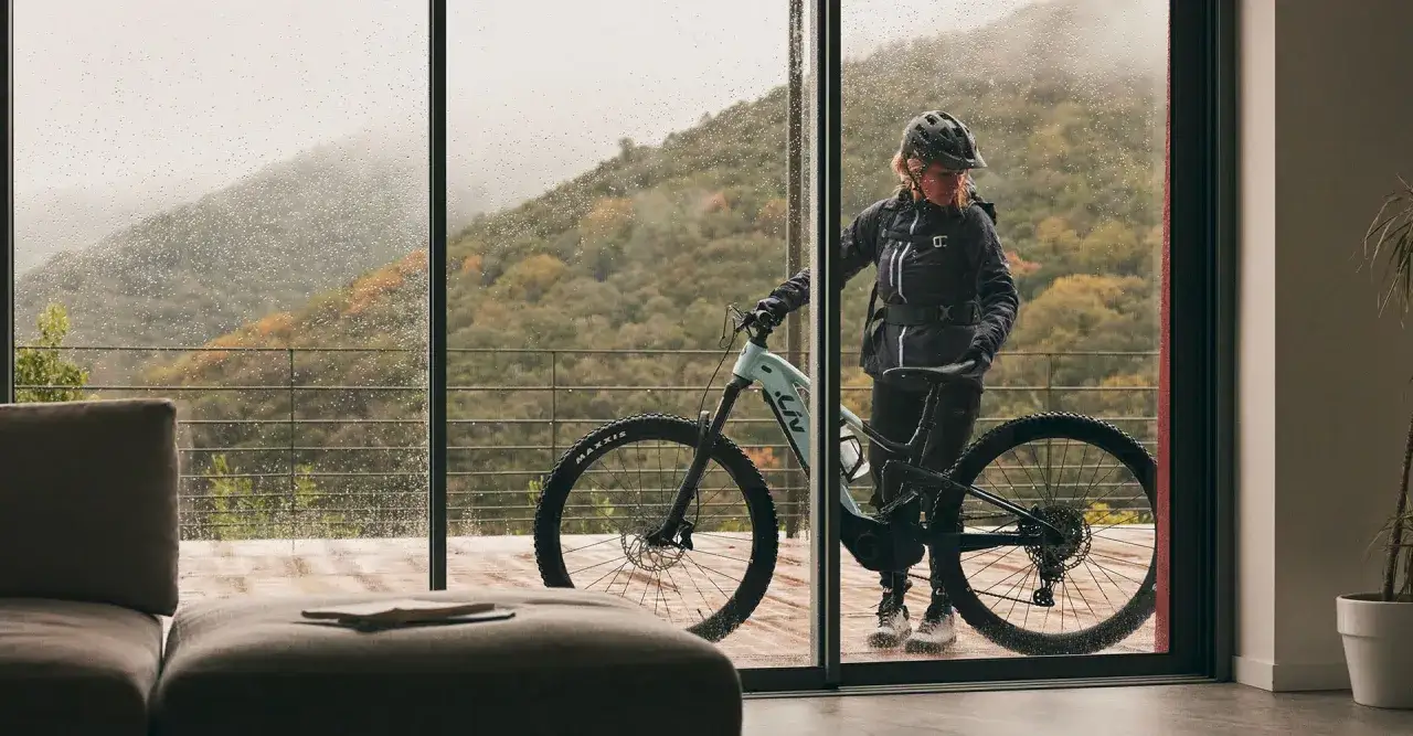Kobieta w kasku i kurtce stoi z rowerem elektrycznym przy drzwiach tarasowych. Za szybą widać deszcz i g&oacute;rzysty krajobraz.