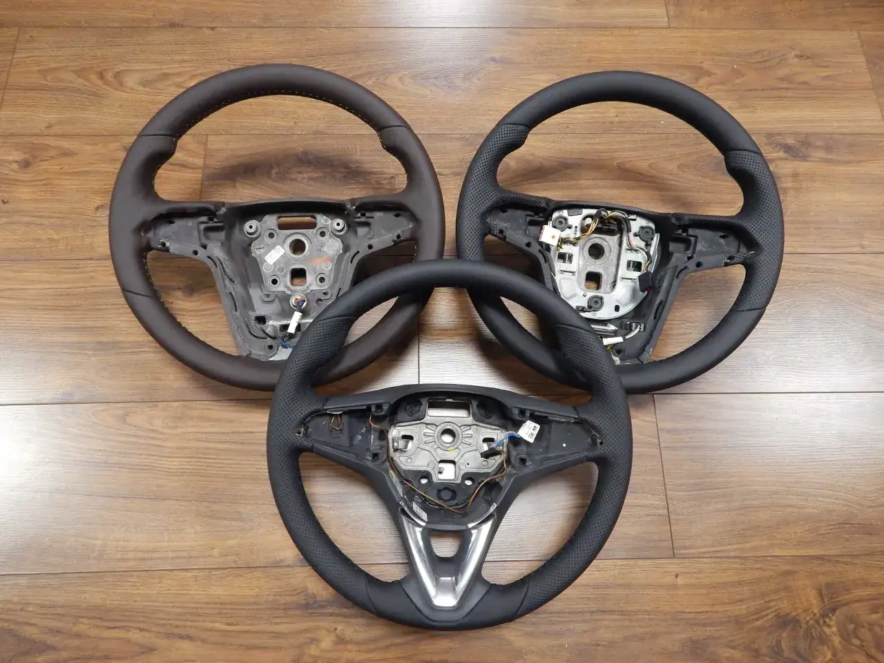 Trzy kierownice Opel Astra J, dwie skórzane z perforacją, jedna gładka, leżą na drewnianej podłodze.