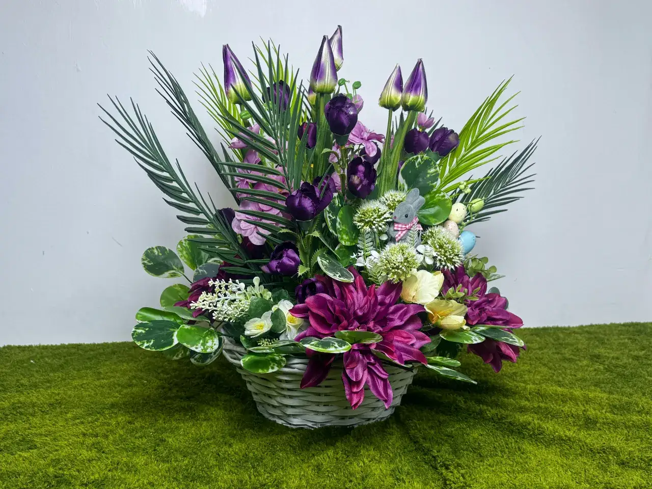 Stroik z żywych kwiatów na cmentarz: kosz pełen fioletowych i różowych kwiatów, zielonych liści, zajączka i pisanek.