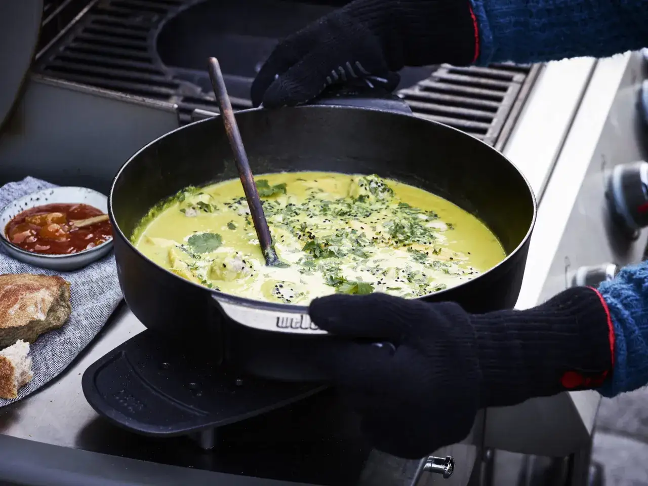 Ręce w rękawicach trzymają żeliwną patelnię z kremową zupą z grilla, posypaną ziołami i czarnym sezamem. Obok chleb i miseczka z sosem.