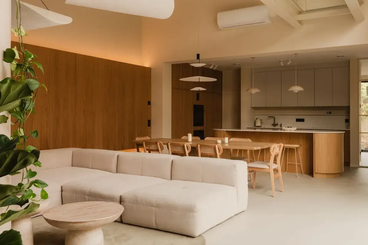 Jasne, przestronne wnętrze z sofą, stołem i aneksem kuchennym. Doświetlenie pomieszczeń światłem naturalnym podkreśla ciepło drewna i jasnych tkanin.