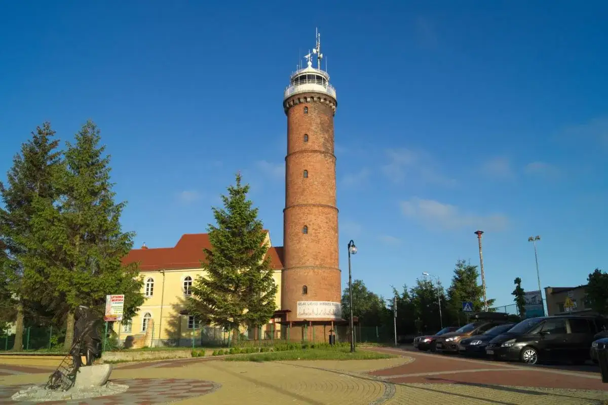 Wysoka, ceglana latarnia morska Jarosławiec góruje nad budynkami i drzewami.