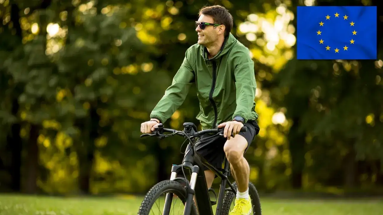 Uśmiechnięty mężczyzna w zielonej kurtce jedzie na rowerze typu s-pedelec po parku. Obok flaga UE.
