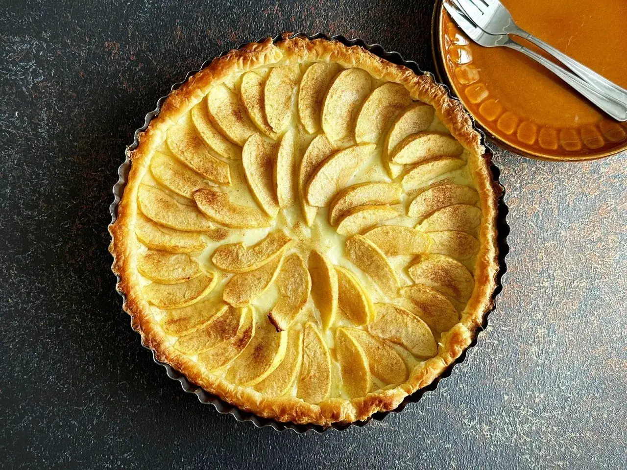 Apetyczna tarta na kruchym spodzie z jabłkami i kremem, posypana cynamonem. Obok talerzyk z widelcem.