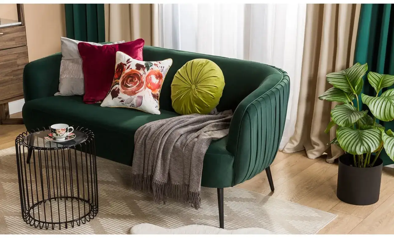 Zielona kanapa z poduszkami i pledem, stolik kawowy z filiżanką, roślina w doniczce &ndash; to wszystko tworzy przytulny, **pok&oacute;j retro**.