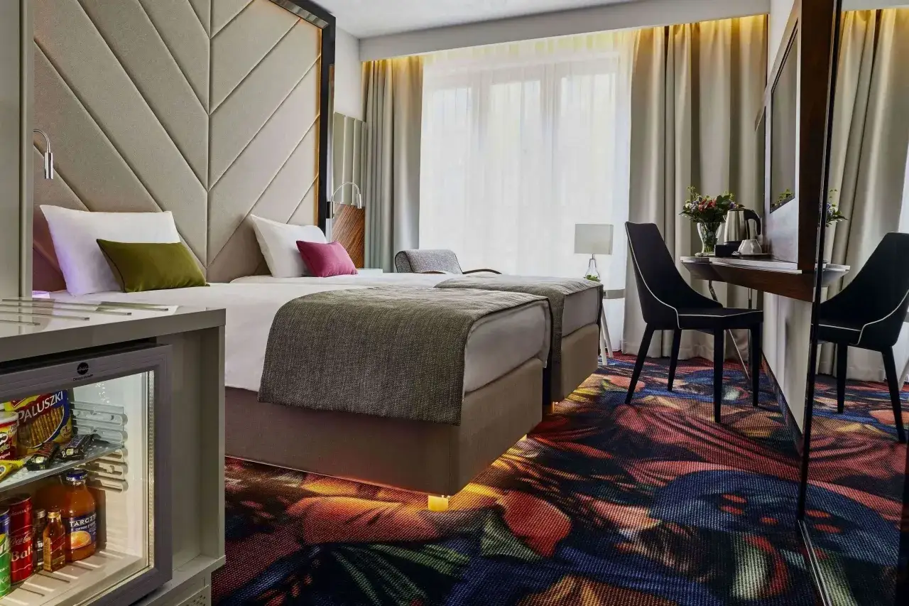 Nowoczesny pokój hotelowy z dwoma łóżkami, minibarem i kolorowym dywanem. Sprawdź, ile kosztuje nocleg w Krakowie.