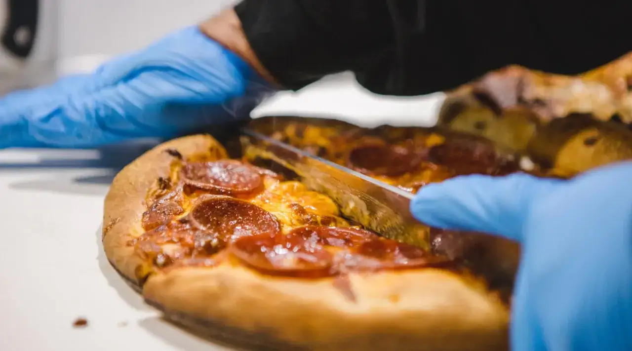 Ręce w niebieskich rękawiczkach kroją pyszną pizzę z grilla z pepperoni.