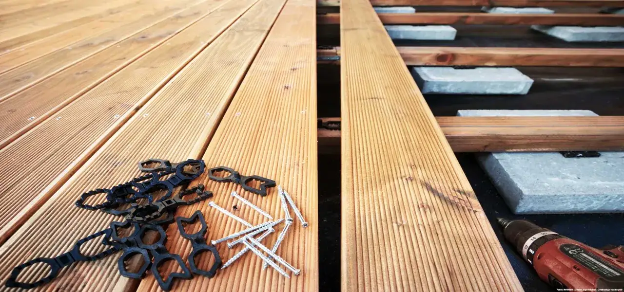 Drewniany taras w budowie. Widać deski, wkręty, klipsy montażowe i wiertarko-wkrętarkę. Zastanawiasz się, ile kosztuje taras drewniany?